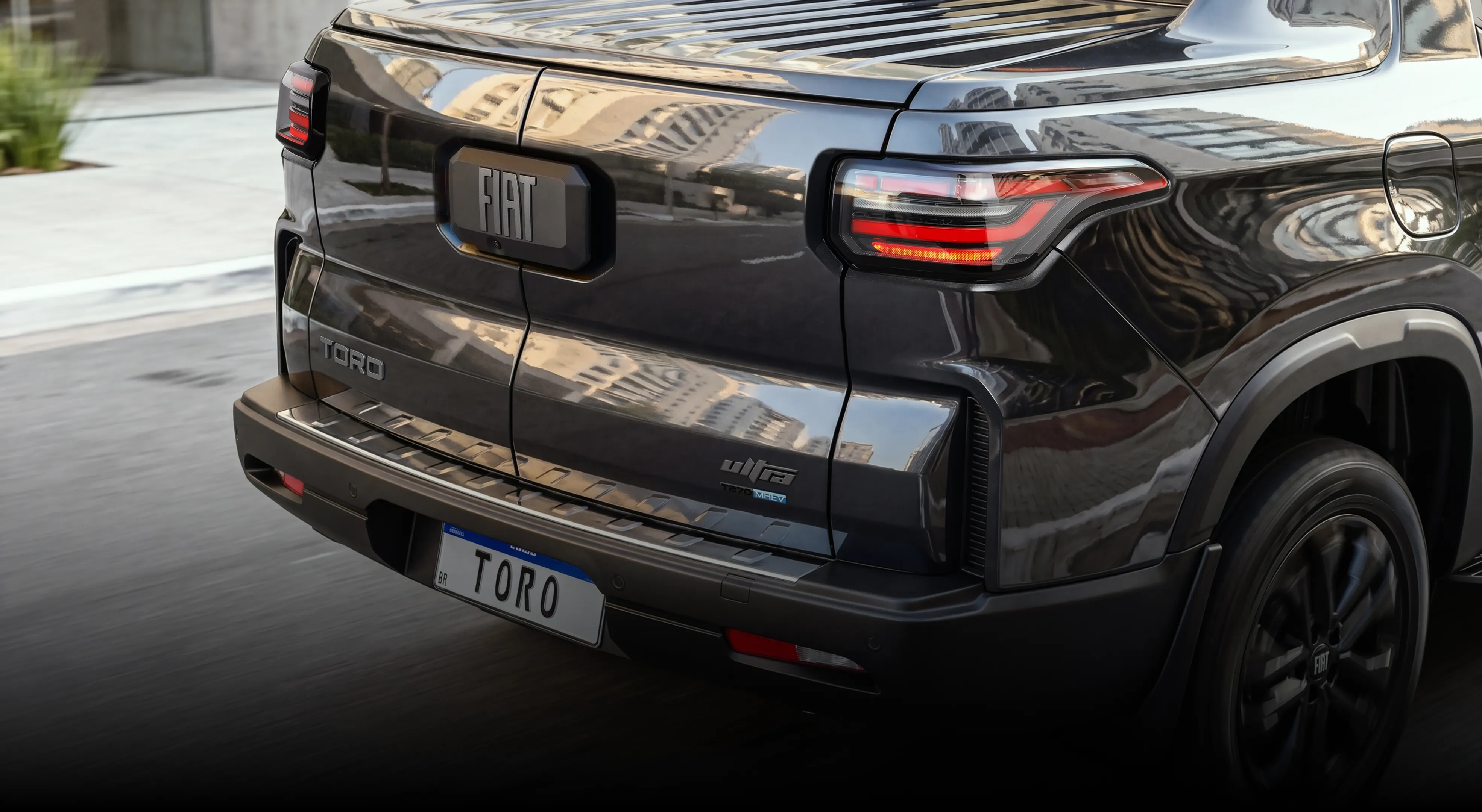 Traseira da Fiat Toro preta em movimento, com destaque para o logo FIAT centralizado na tampa da caçamba, lanternas acesas em LED e identificação do modelo “Toro”. O para-choque tem degrau integrado e, na placa do veículo, lê-se ”TORO”.