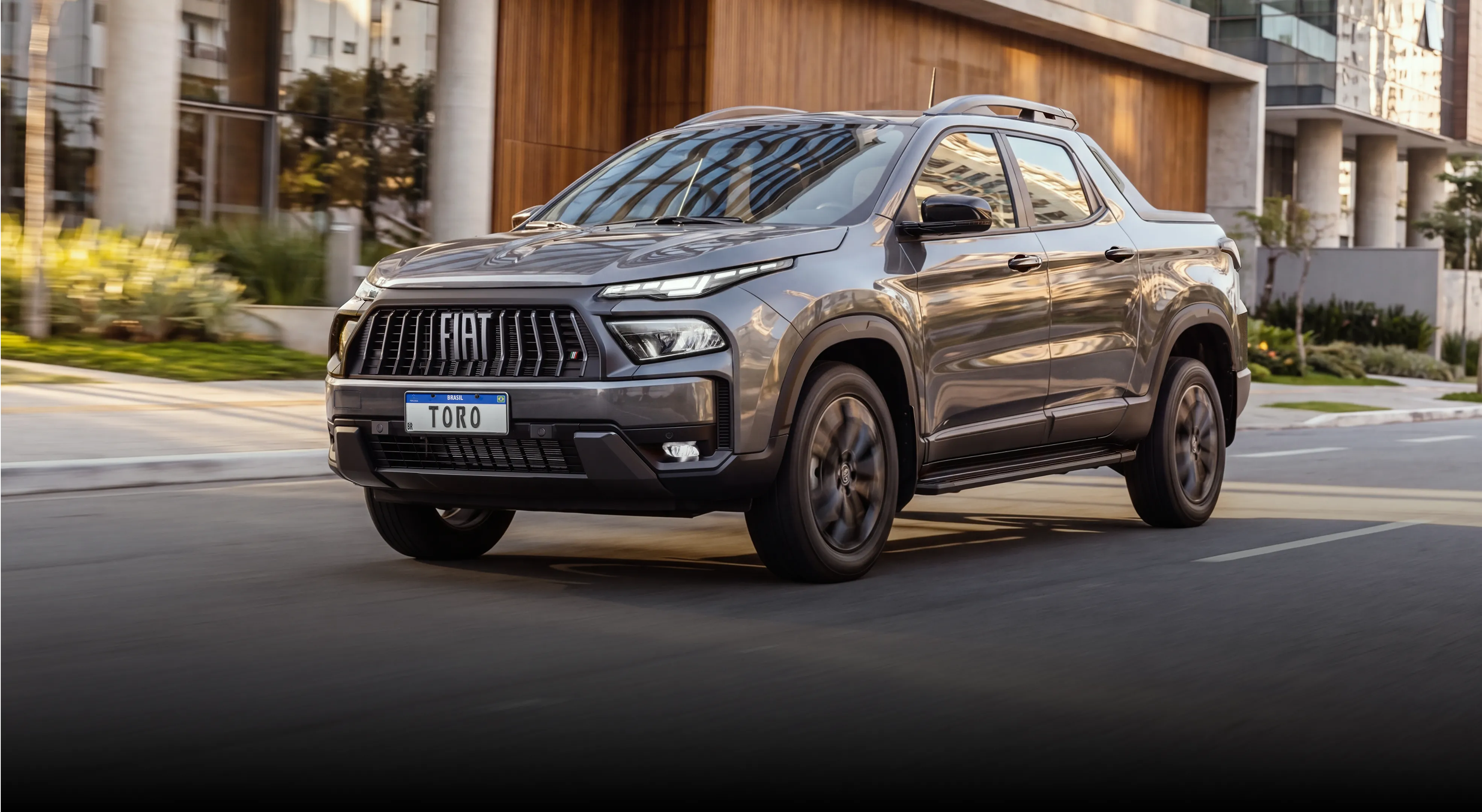 Picape Fiat Toro cinza em movimento em via urbana, com destaque para a grade frontal com o logo FIAT, faróis acesos e carroceria robusta, com prédios modernos e vegetação ao fundo.