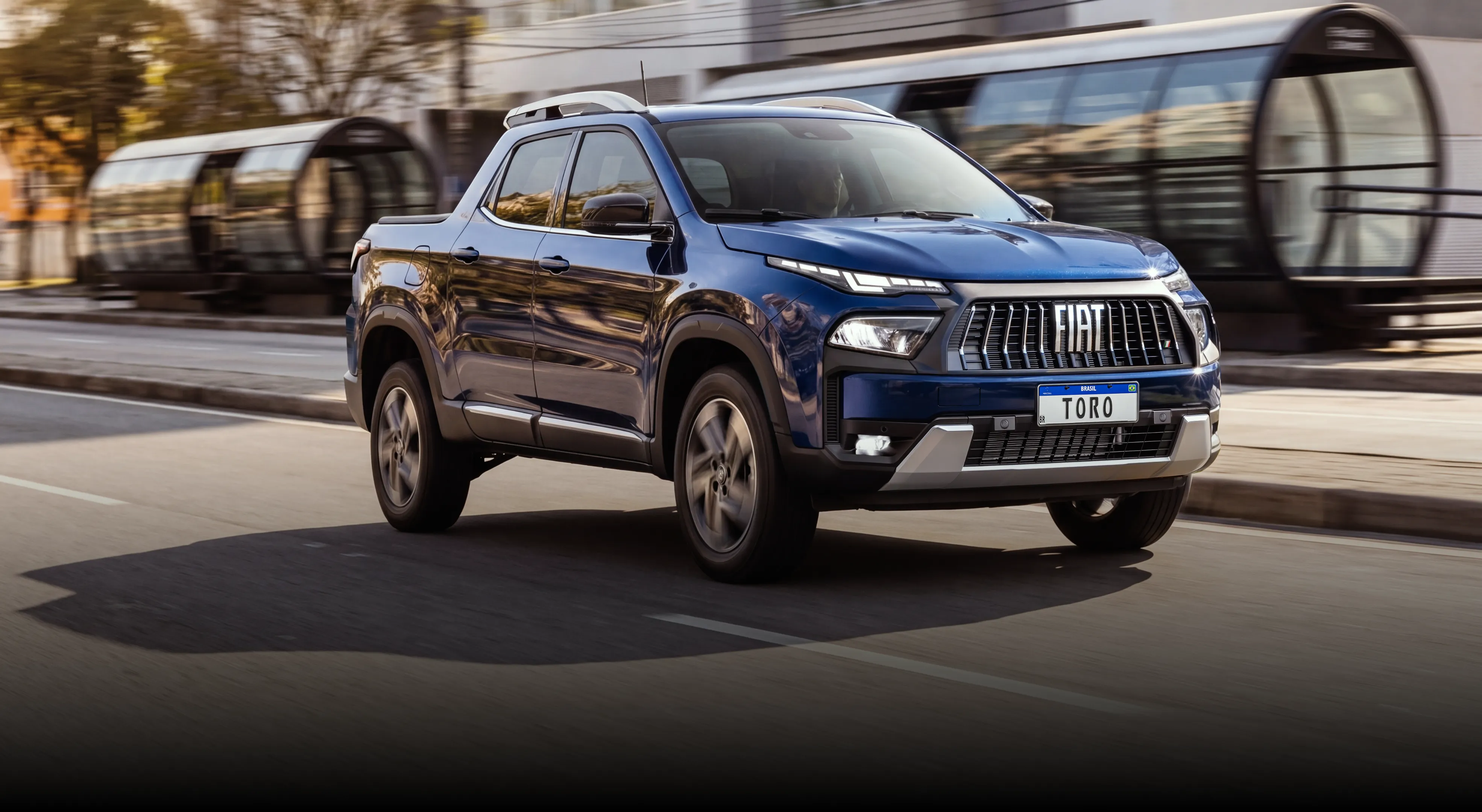 Picape Fiat Toro azul em movimento em via urbana, com destaque para a grade frontal com o logo FIAT e faróis acesos. Ao fundo, estação de transporte com estruturas de vidro e cenário levemente desfocado indicam velocidade.