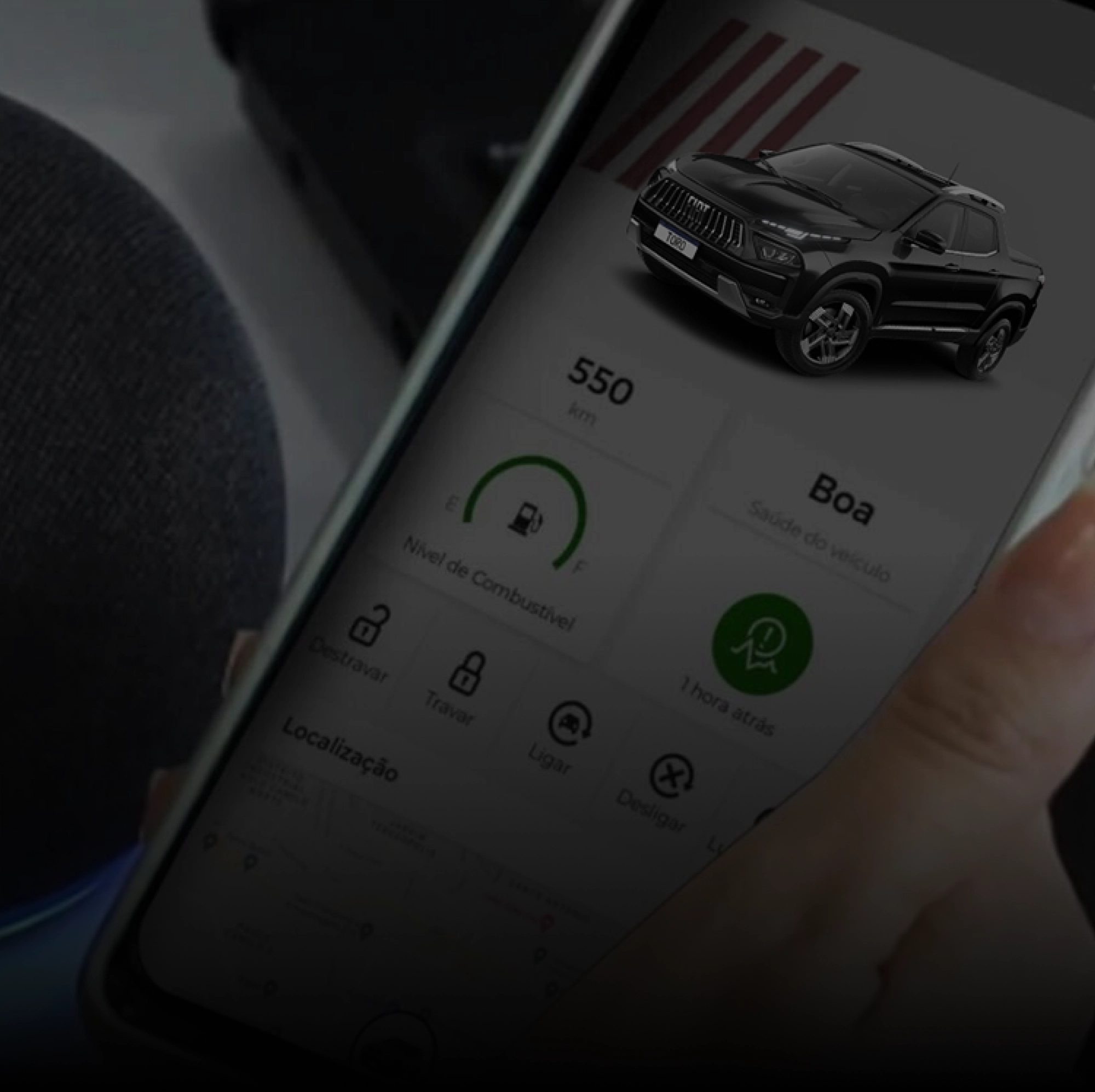 Uma mão segura um smartphone com aplicativo aberto exibindo controles do veículo. Ao fundo, há uma Fiat Toro preta.